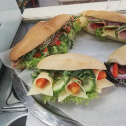 Destin Anna Pajnowska - Cztery bagietki z różnymi nadzieniami: szynką, serem, warzywami i kiełkami, ułożone na papierze na srebrnej tacy.