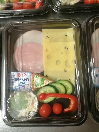 Plastikowe pudełko z gotową przekąską: wędlina, ser żółty z dziurami, serek topiony, kostka masła, sałatka jarzynowa, ogórek, papryka i pomidorki koktajlowe.