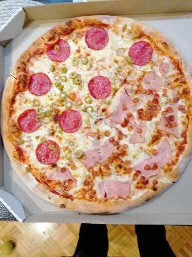 Pizza w kartonie z dwoma różnymi połówkami: jedna z salami i oliwkami, druga z szynką i małymi kiełbaskami.