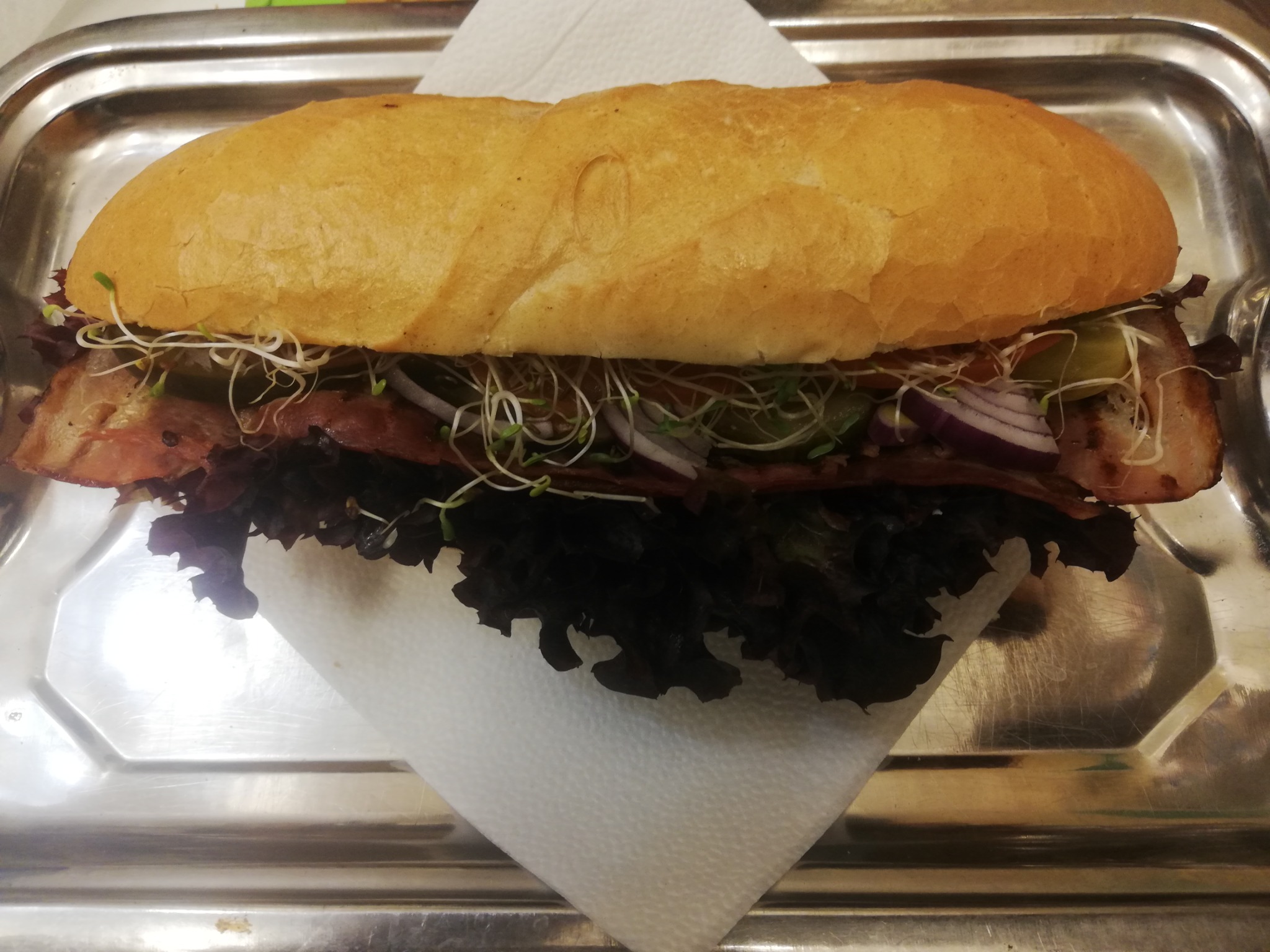 Podłużna kanapka z boczkiem, sałatą, czerwoną cebulą i kiełkami, ułożona na serwetce na metalowej tacy.