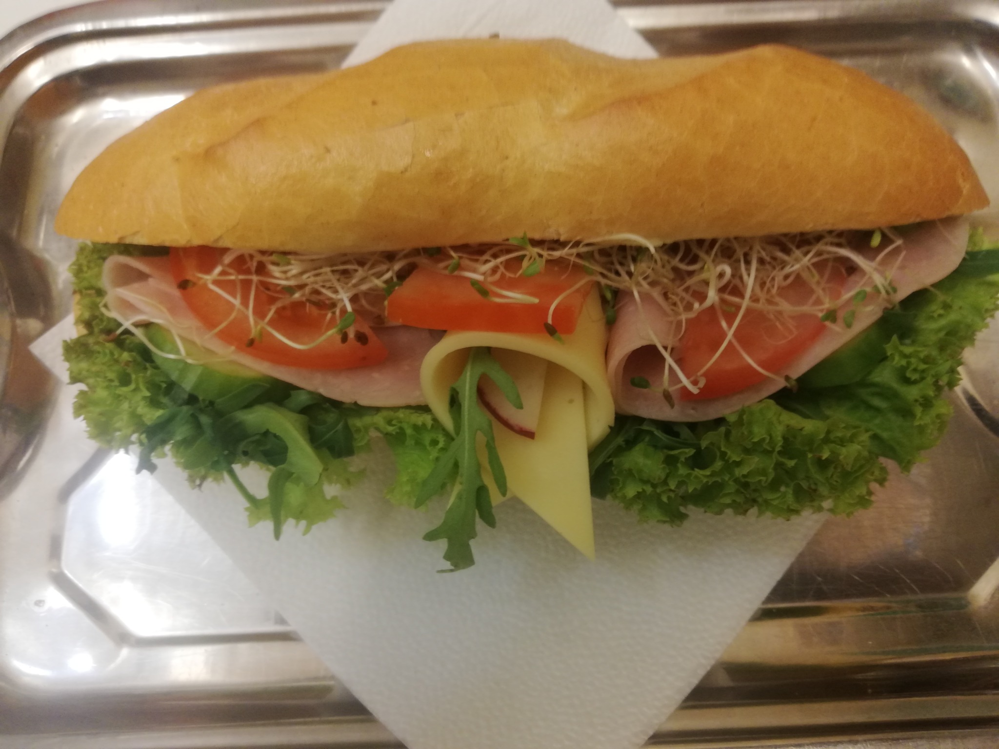 Świeża bagietka z szynką, serem, pomidorem, sałatą i kiełkami, podana na srebrnej tacy na białej serwetce.