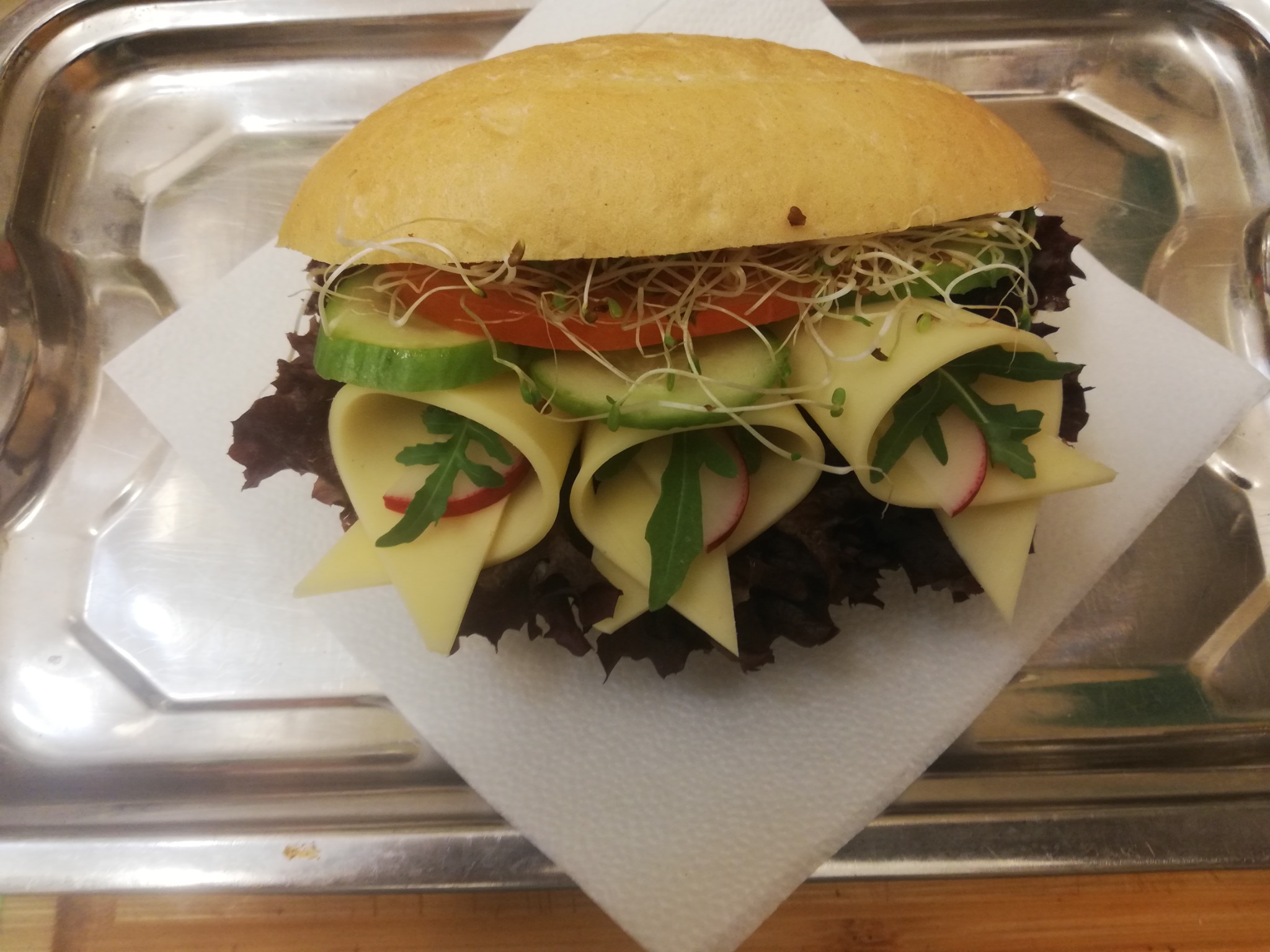 Kanapka z serem, rzodkiewką, ogórkiem, pomidorem i kiełkami na serwetce i metalowej tacy.