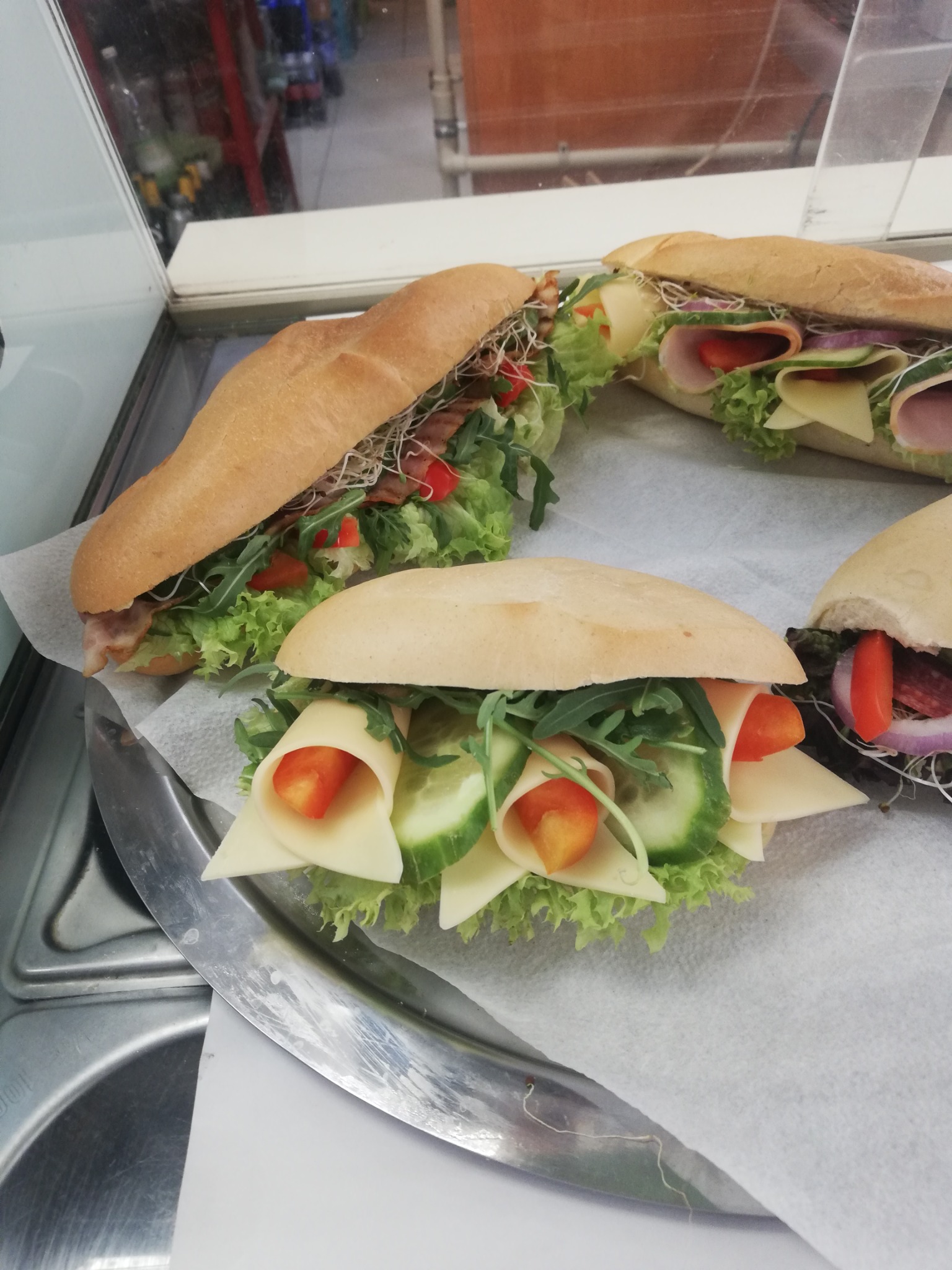 Cztery bagietki z różnymi nadzieniami: szynką, serem, warzywami i kiełkami, ułożone na papierze na srebrnej tacy.