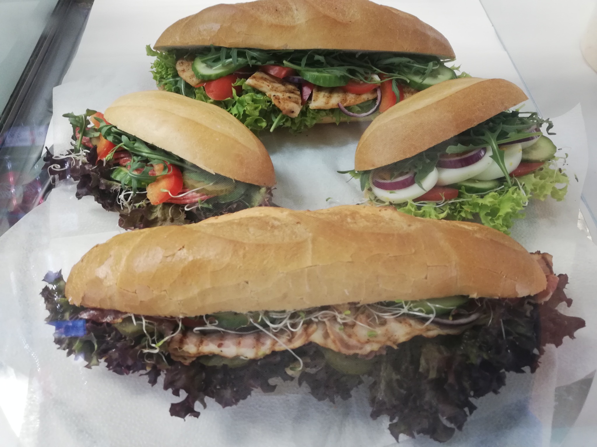 Cztery świeże bagietki z grillowanym kurczakiem, pomidorami, ogórkiem, czerwoną cebulą, rukolą i sałatą, ułożone na białym papierze.