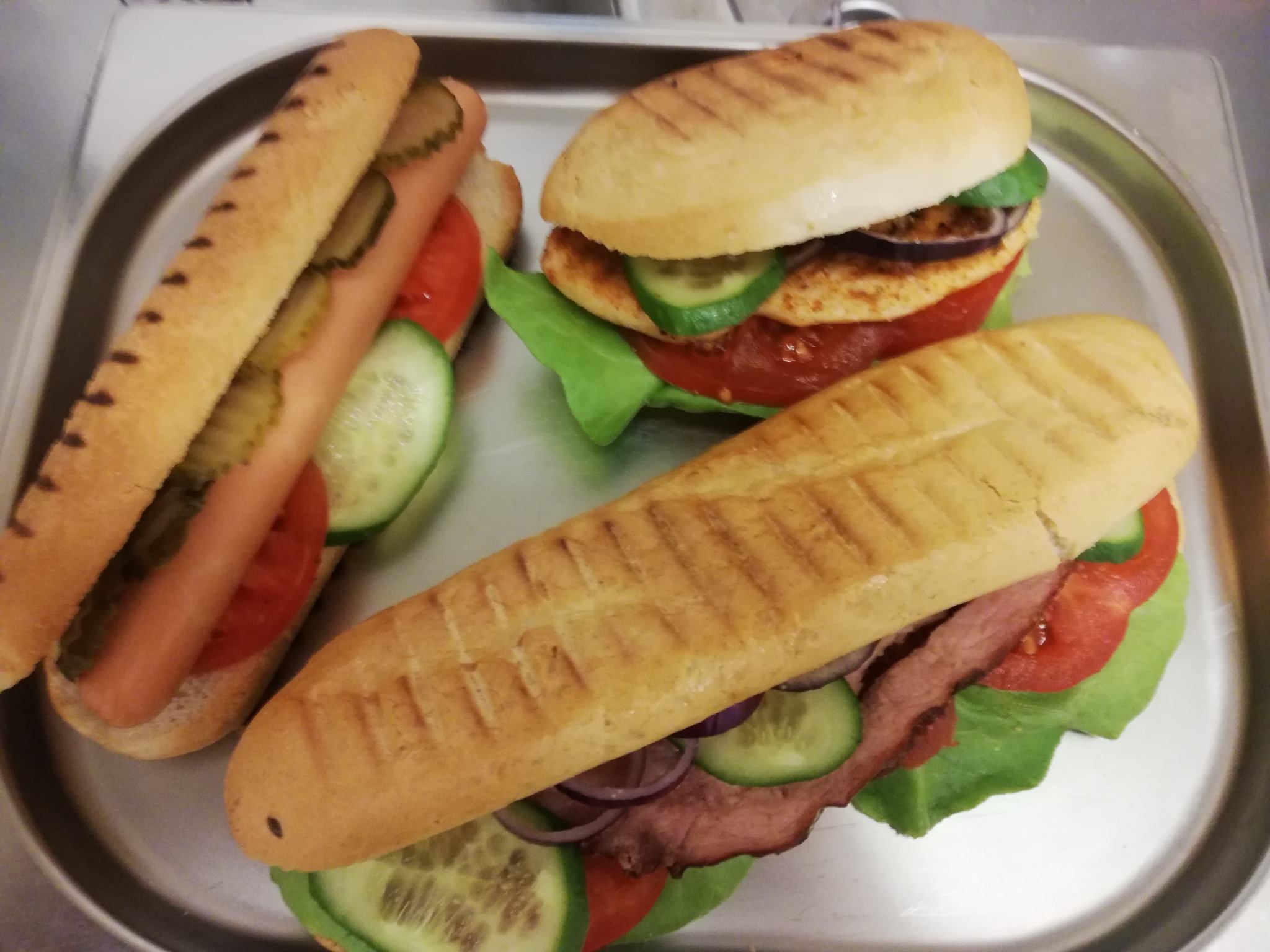 Trzy różne kanapki na srebrnej tacy: hot dog z ogórkiem konserwowym, kanapka z kurczakiem i warzywami oraz kanapka z wędliną, pomidorem, ogórkiem i czerwoną cebulą.