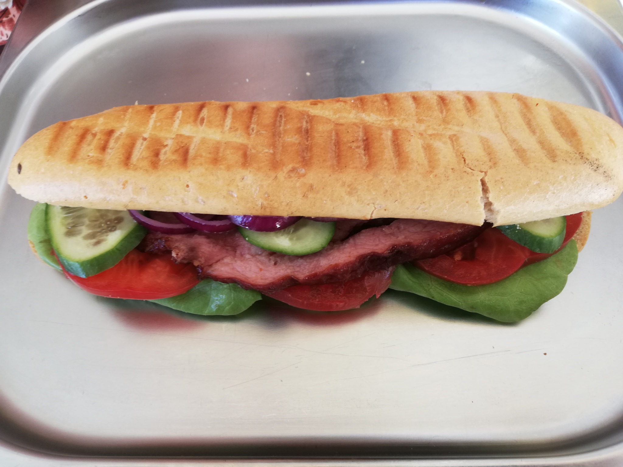 Podłużna kanapka z grillowaną bułką, wołowiną, sałatą, pomidorem, ogórkiem i czerwoną cebulą, ułożona na metalowej tacy.