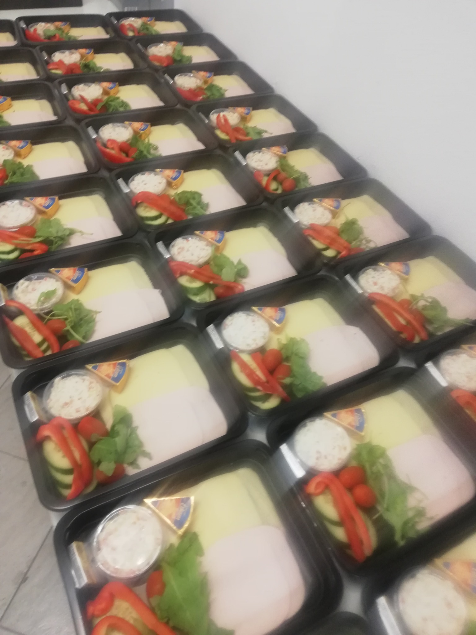 Rzędy czarnych pojemników z gotowymi zestawami kanapek: ser, szynka, warzywa i dip, przygotowane do transportu.