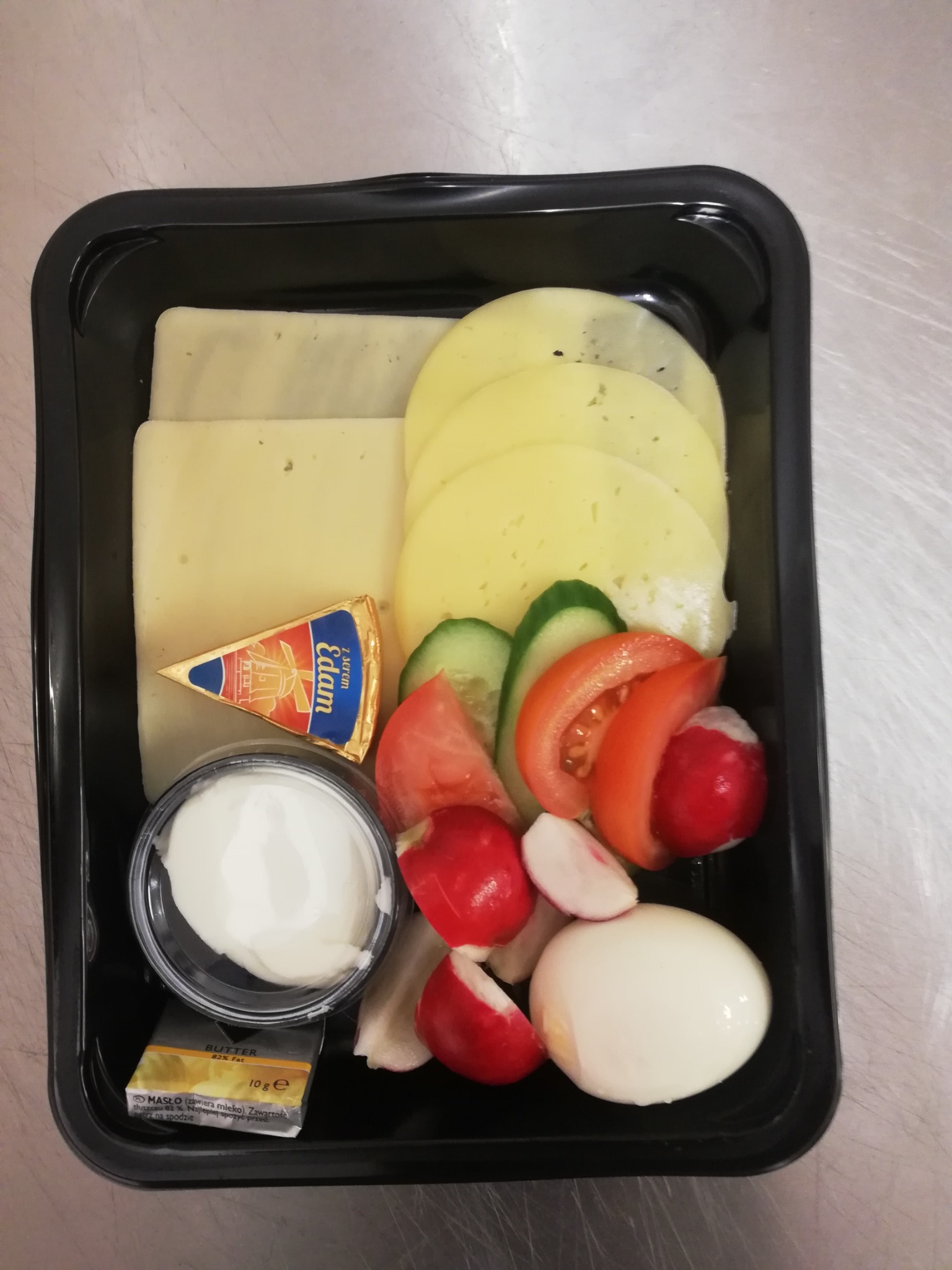Czarna tacka z przekąskami: plastry sera żółtego, trójkątny serek Edam, pojemnik z sosem, kostka masła, plasterki ogórka i pomidora, rzodkiewki i jajko na twardo.