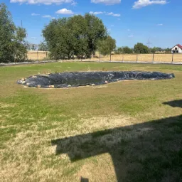 Wykopany staw ogrodowy z czarną folią, otoczony kamieniami, na trawniku w słoneczny dzień.