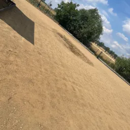 Przygotowany teren pod trawnik z widocznymi śladami maszyn, ogrodzenie w tle i drzewa w oddali pod błękitnym niebem.