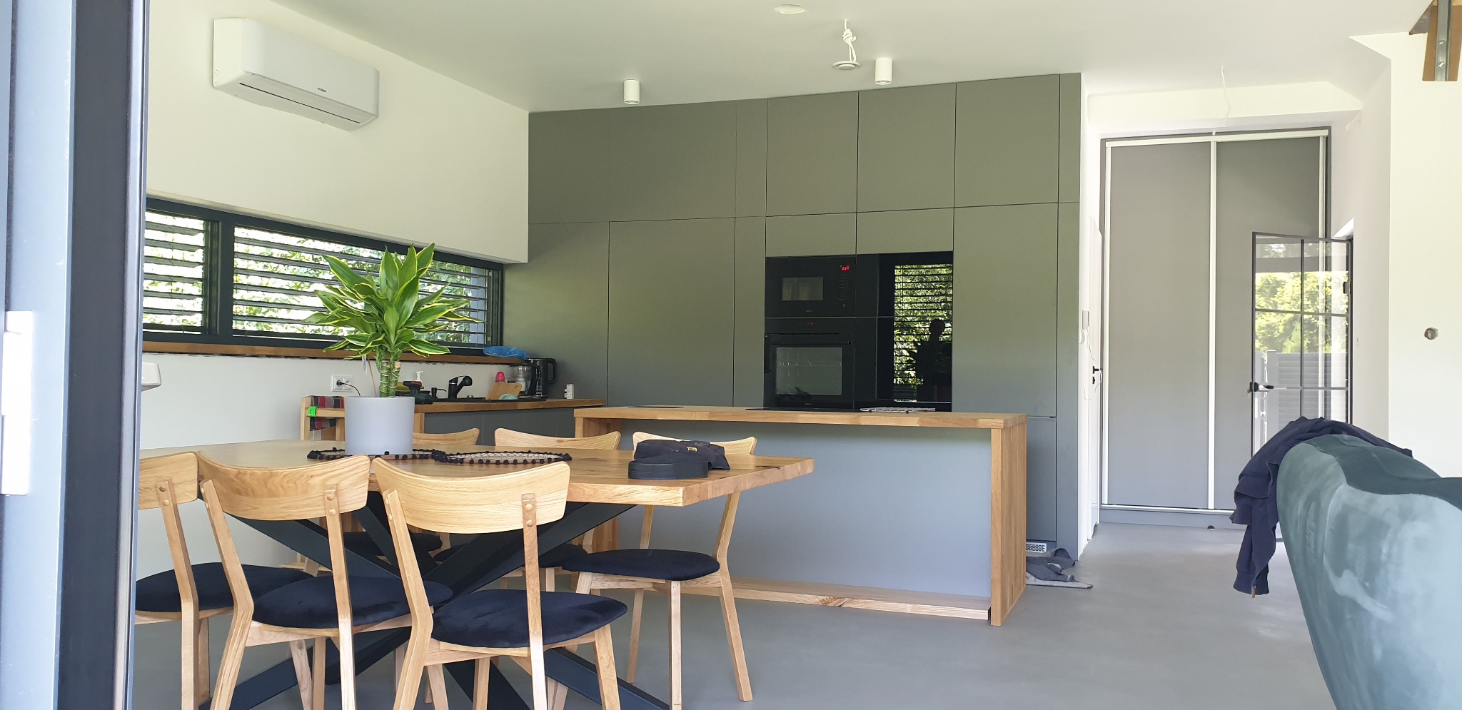 Nowoczesna kuchnia z szarymi frontami szafek, drewnianym blatem i wyspą; stół z drewnianym blatem i krzesłami z czarnymi siedziskami, roślina doniczkowa na stole, okno z żaluzjami w tle.