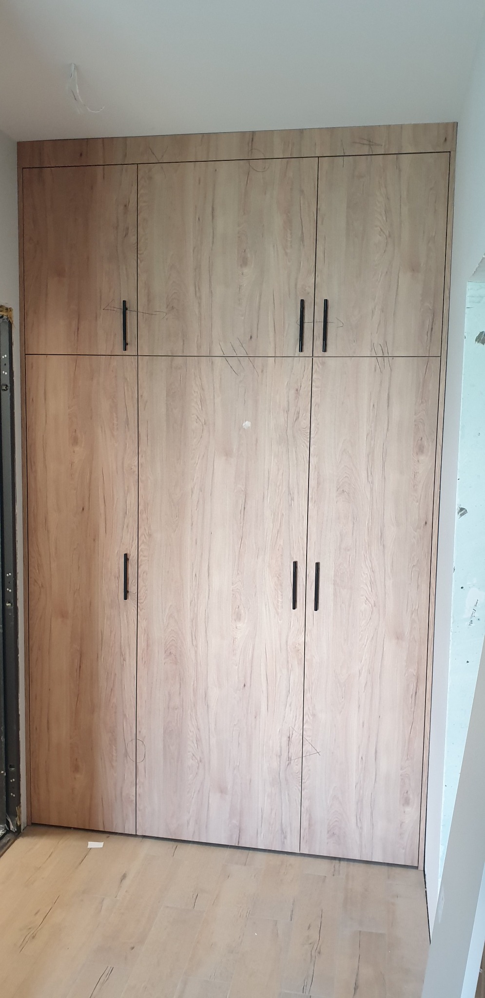 Wysoka zabudowa szafy z frontami w kolorze jasnego drewna, czarne, smukłe uchwyty, widoczne ślady ołówka na frontach, podłoga imitująca drewno, wnętrze w trakcie wykańczania.
