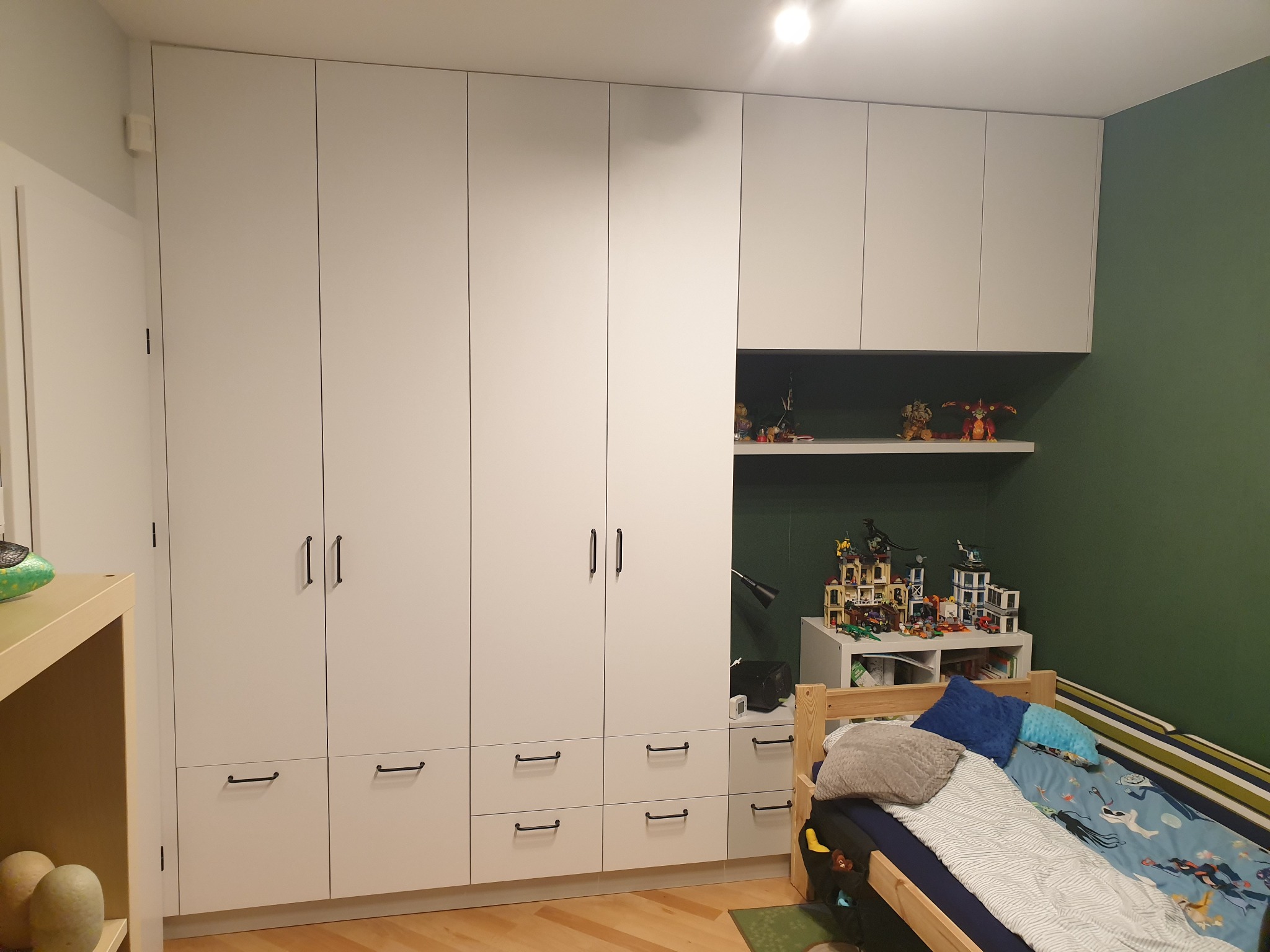 Biała szafa wnękowa z czarnymi, podłużnymi uchwytami w pokoju dziecięcym, z półkami wypełnionymi zabawkami i łóżkiem z pościelą w superbohaterów.