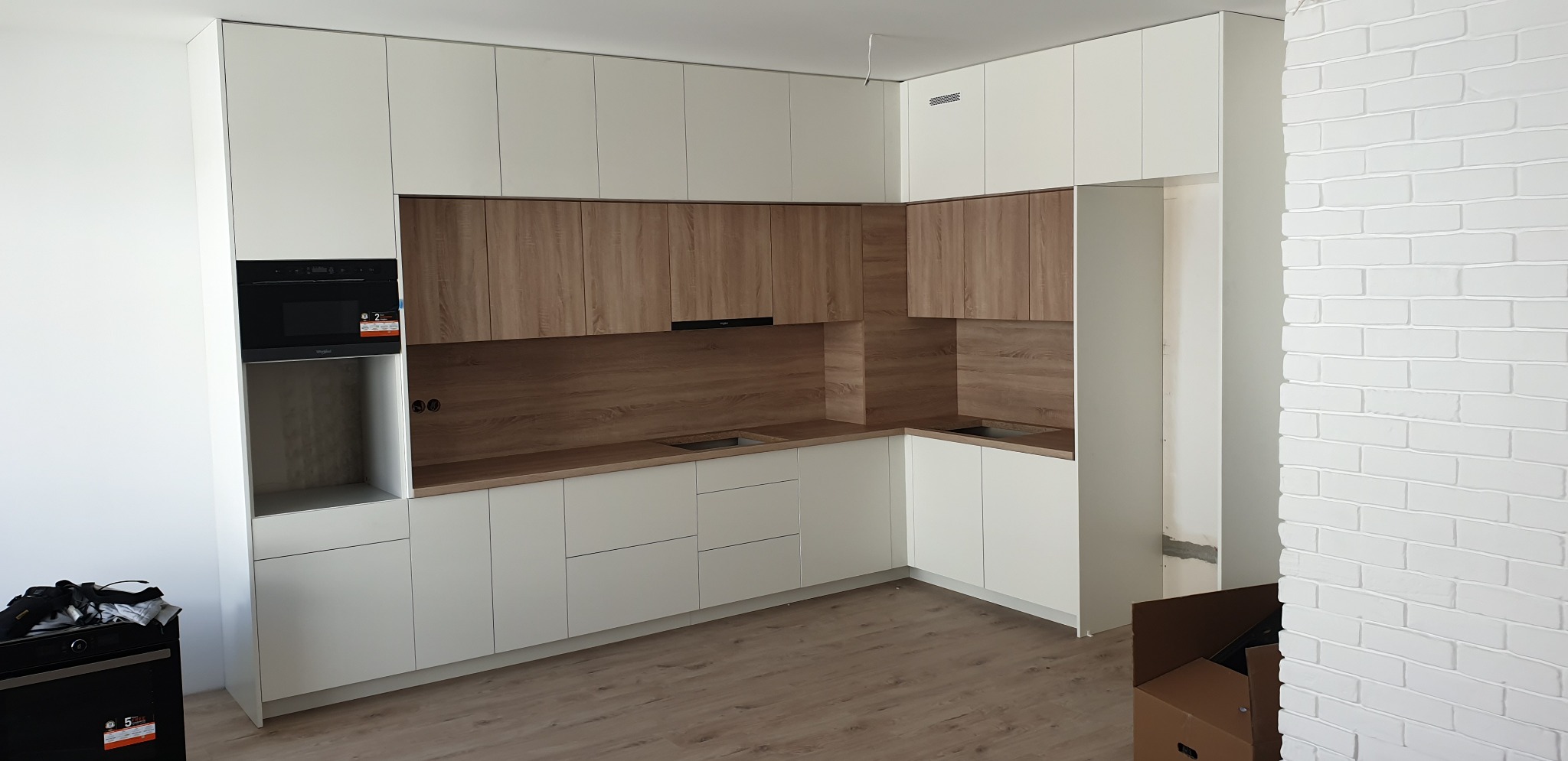 Jasna kuchnia z drewnianymi akcentami, zabudowa pod sufit, widoczny piekarnik i fragment ściany z białej cegły.
