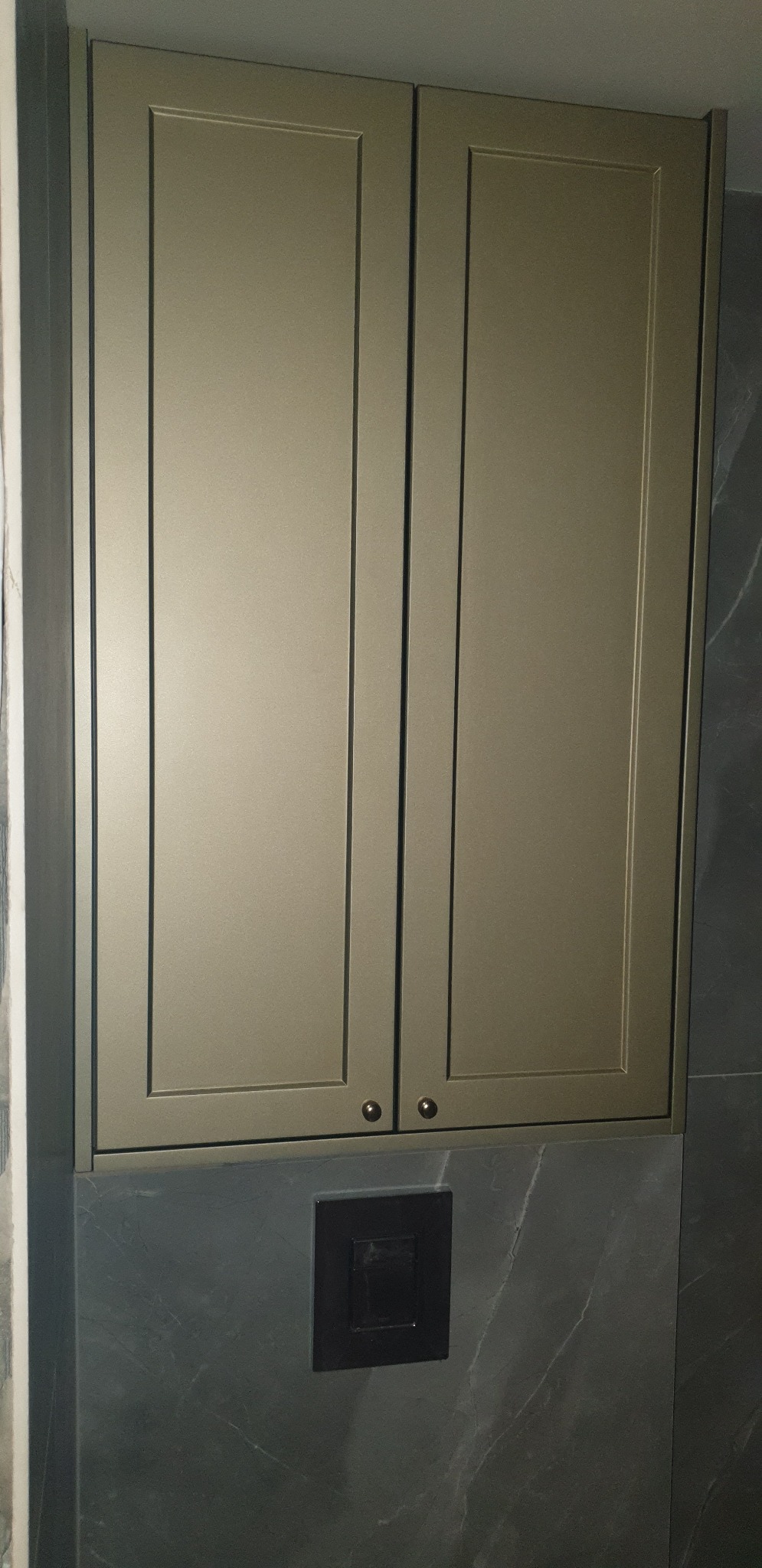 Zamknięta szafka łazienkowa w kolorze oliwkowym, z dwoma drzwiczkami i okrągłymi uchwytami, zamontowana nad spłuczką wbudowaną w ścianę wykończoną szarym marmurem.