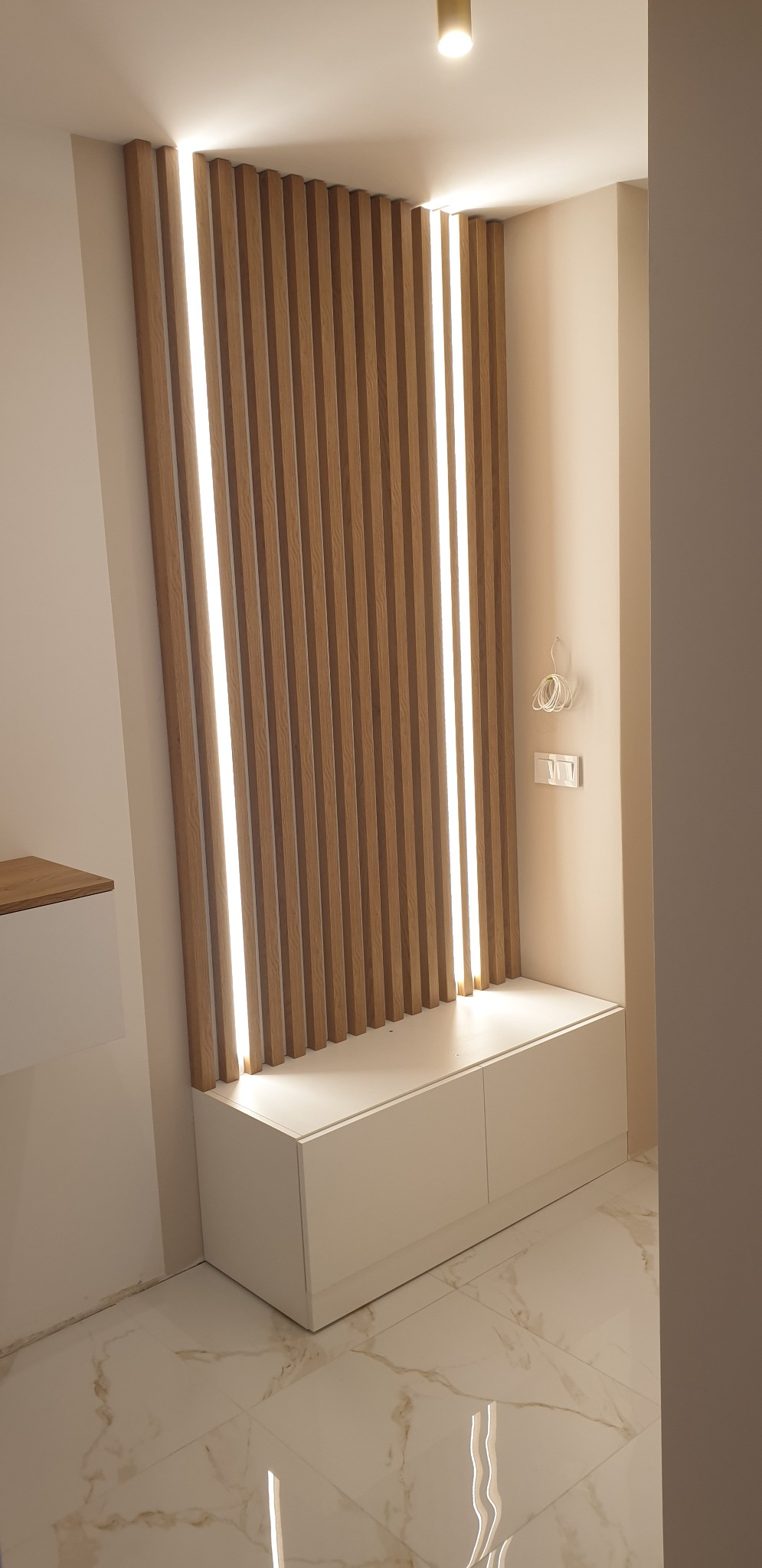 Nowoczesna aranżacja przedpokoju z drewnianymi lamelami podświetlonymi ledami, białą szafką z dwoma frontami i marmurową podłogą.