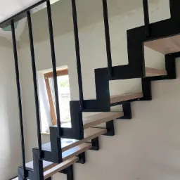 Nowoczesne schody metalowe z drewnianymi stopniami i czarną, minimalistyczną konstrukcją, widoczne na tle ściany w trakcie wykończenia i okna.