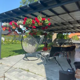 Zadaszona przestrzeń rekreacyjna z metalową konstrukcją dachu, wiszącymi donicami z czerwonymi i białymi kwiatami, wiszącym fotelem, stołem z krzesłami i kosiarką.