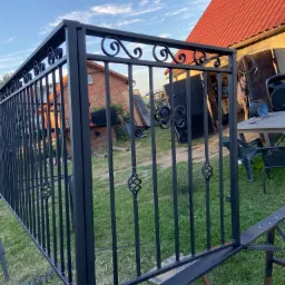 Fragment czarnego ogrodzenia metalowego z ozdobnymi elementami, widoczne elementy montażowe, w tle dom z czerwoną dachówką i niebieskie niebo.