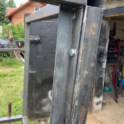 Surowa, metalowa konstrukcja w trakcie spawania, widoczne spawy, śruby mocujące i ślady korozji, na tle budynku z czerwonej cegły i częściowo widocznego warsztatu.