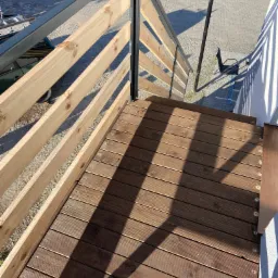 Widok z góry na drewniany taras z jasną, drewnianą balustradą z metalowymi słupkami, rzucającą cienie na deski tarasowe; w tle widoczny fragment zielonego samochodu i zabawka dziecięca.