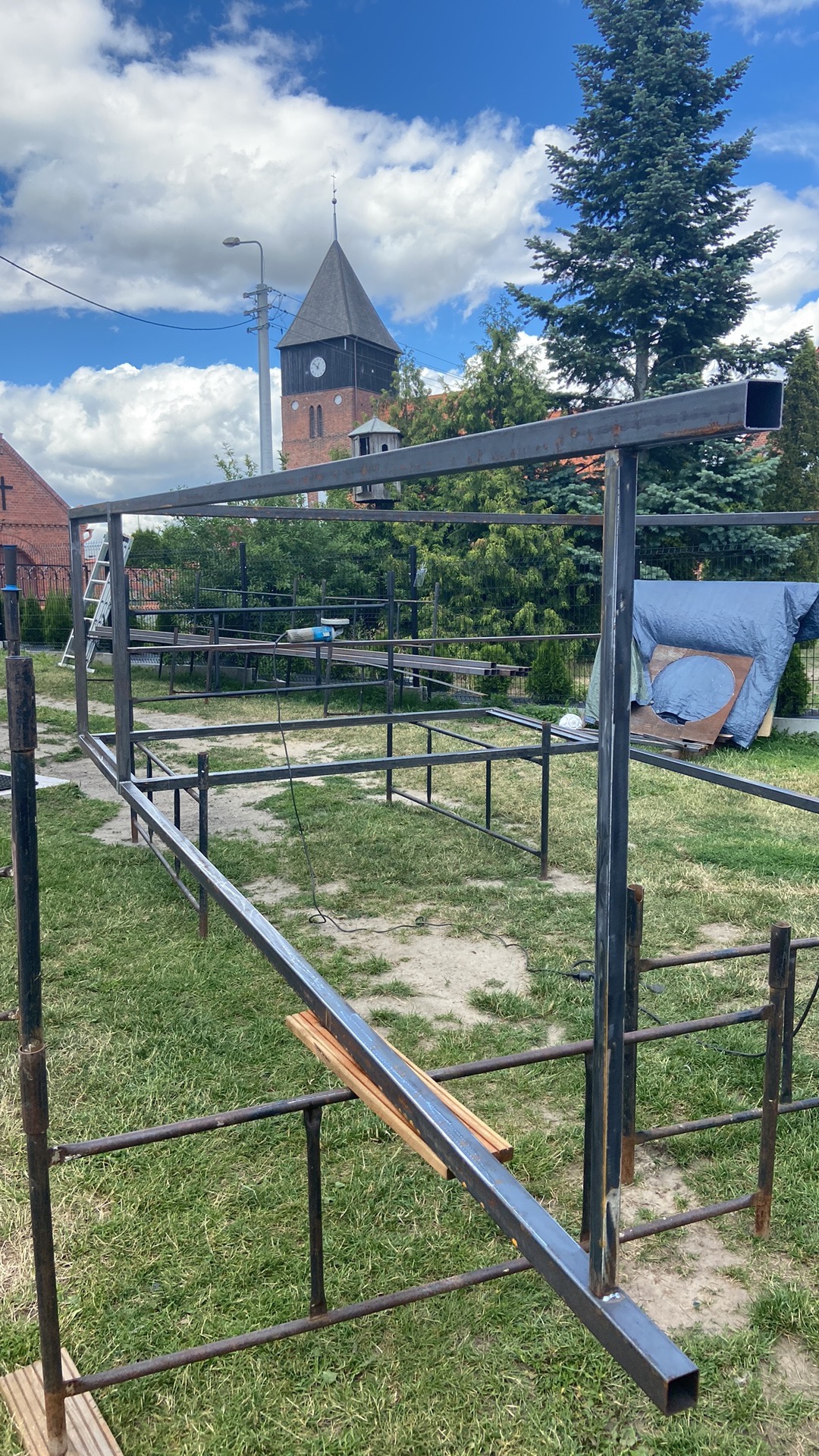 Konstrukcja metalowa w trakcie budowy na trawniku, z widocznym kościołem w tle, elementy spawane z profili kwadratowych.