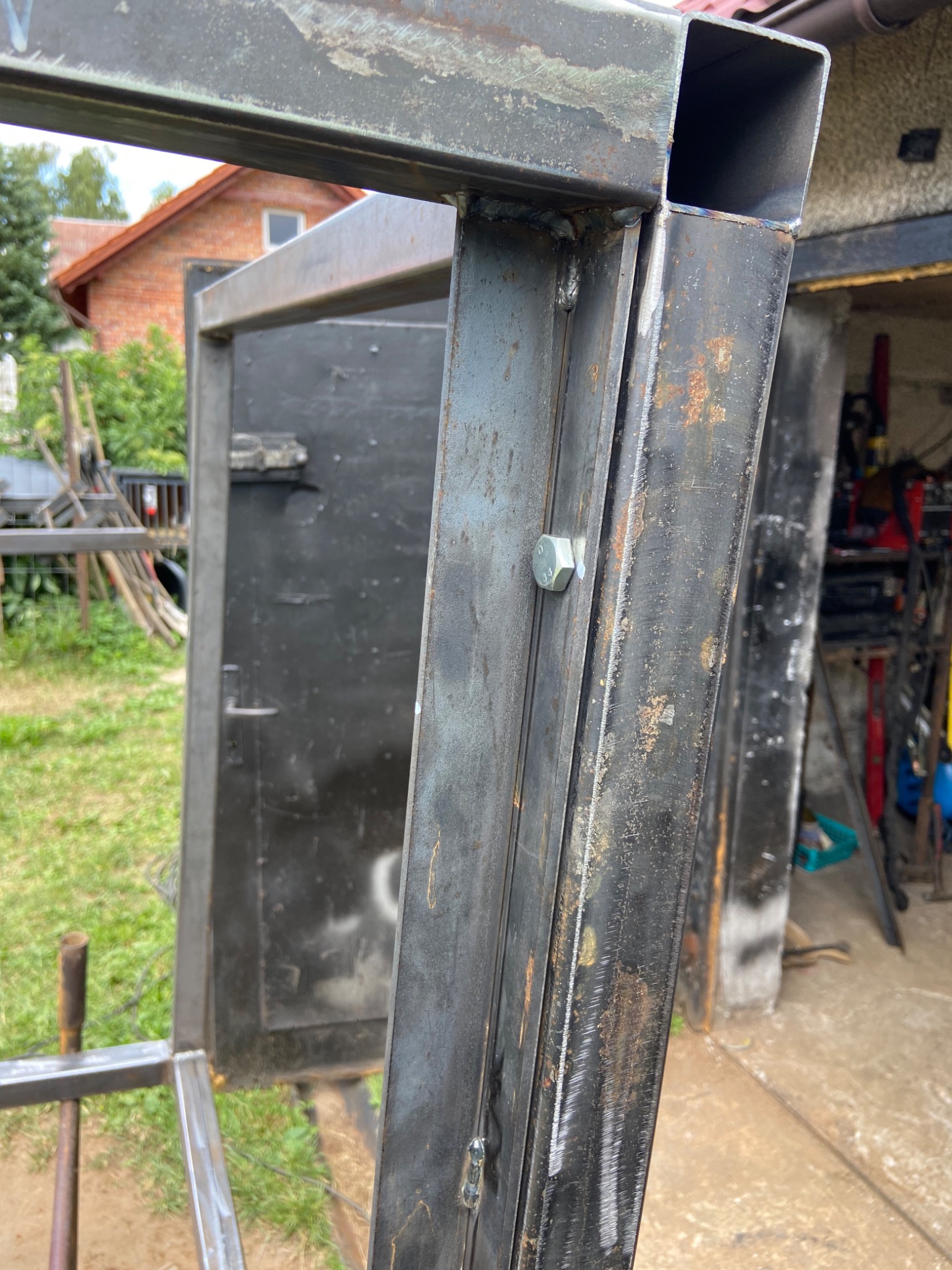 Surowa, metalowa konstrukcja w trakcie spawania, widoczne spawy, śruby mocujące i ślady korozji, na tle budynku z czerwonej cegły i częściowo widocznego warsztatu.