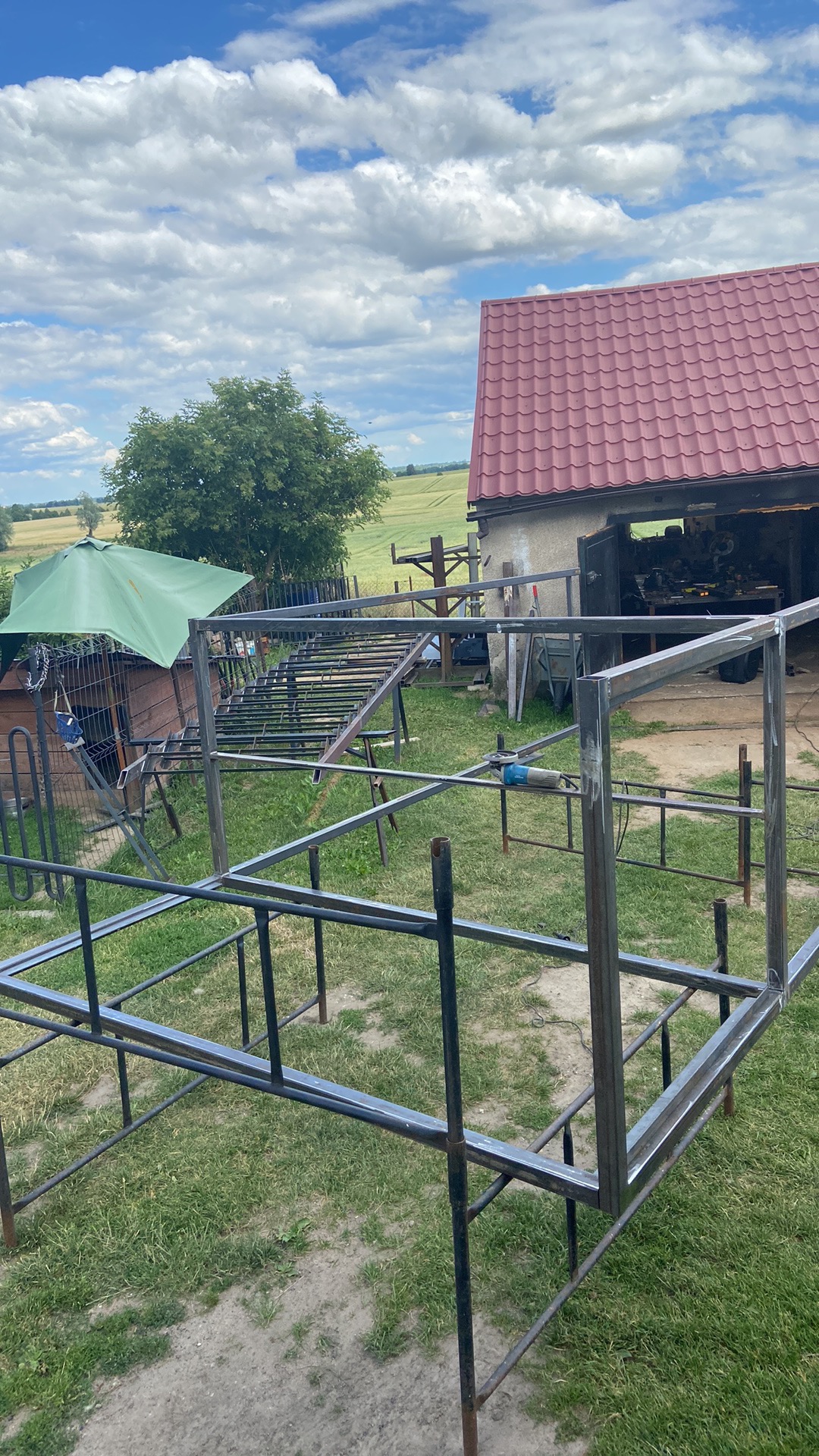 Metalowa konstrukcja szkieletowa na trawie, prawdopodobnie w trakcie budowy, widoczny warsztat w tle i krajobraz wiejski.