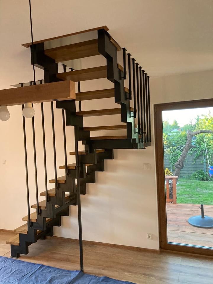 Schody wewnętrzne metalowe z drewnianymi stopniami, czarna konstrukcja, widok z dołu, okno z widokiem na ogród w tle.