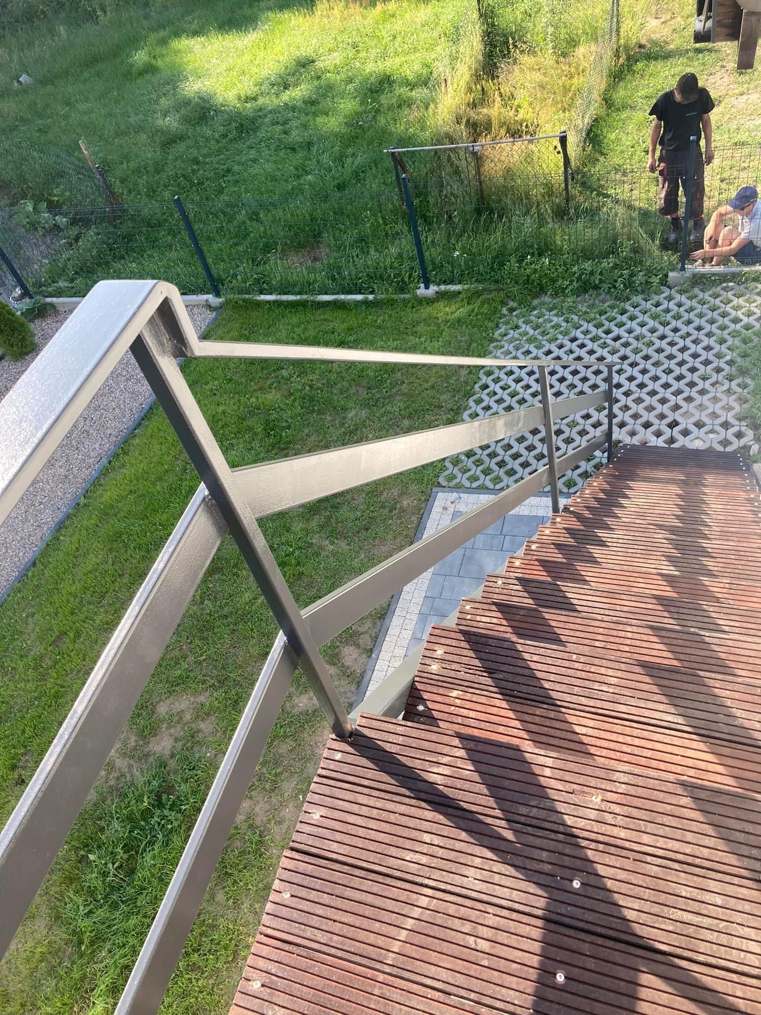 Zewnętrzne schody z drewnianymi stopniami i metalową, pomalowaną na szaro balustradą, widok z góry na ogród z elementami ogrodzenia panelowego w tle.