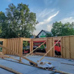 Wznoszenie szkieletu drewnianej konstrukcji budynku na betonowej płycie, widoczny czerwony dźwig za ścianą i budynek apteki w tle.