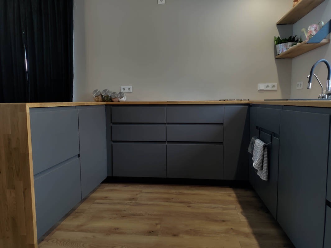 Nowoczesna kuchnia w odcieniach szarości z drewnianym blatem i podłogą, minimalistyczny design, widoczne słoiki z przyprawami na blacie i półka z roślinami.