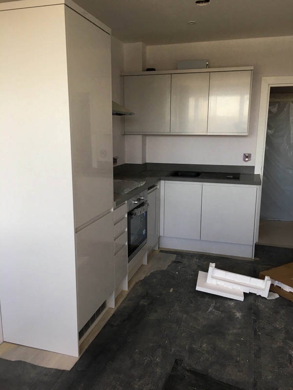 Jasna kuchnia z białymi, lakierowanymi frontami, ciemnym blatem i częściowo położoną podłogą, widoczne elementy montażowe na podłodze.