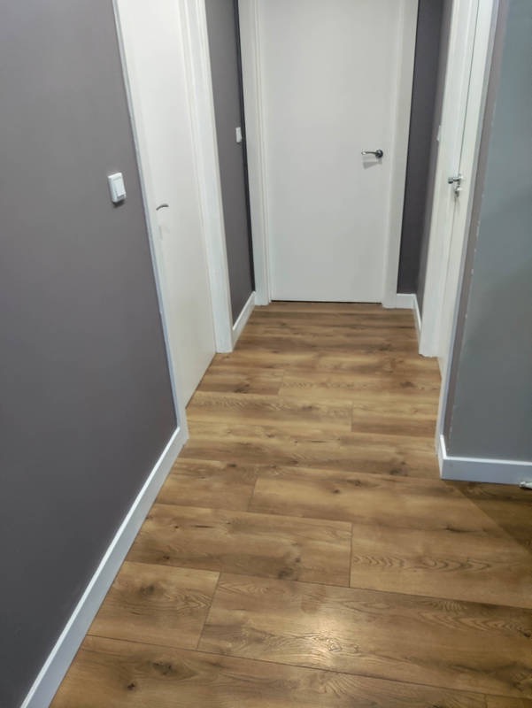 Korytarz z panelami podłogowymi w odcieniach brązu, szare ściany z białymi listwami przypodłogowymi, trzy białe drzwi z srebrnymi klamkami.