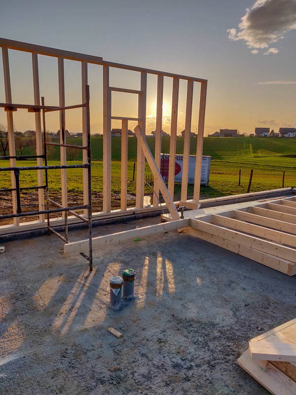 Wczesny etap budowy szkieletu drewnianego domu na betonowej płycie fundamentowej, z widocznymi elementami konstrukcyjnymi i rusztowaniem na tle zachodzącego słońca i zielonych pól.