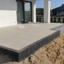 Jasnoszara, kwadratowa płyta tarasowa z ciemnymi obrzeżami z kostki, częściowo ułożona na piasku przy nowoczesnym domu z przesuwnymi drzwiami, widoczne prace wykończeniowe.