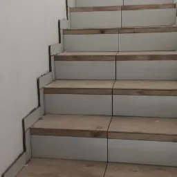 Schody w trakcie wykańczania z płytkami imitującymi drewno i białymi podstopnicami, widoczne krzyżyki dystansowe.