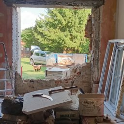Bartosz Naszewski - Wnętrze w trakcie demontażu okna, widoczne gruz, narzędzia, drabina i nowe okno czekające na montaż, na zewnątrz samochód z otwartym bagażnikiem i kontener na odpady budowlane.
