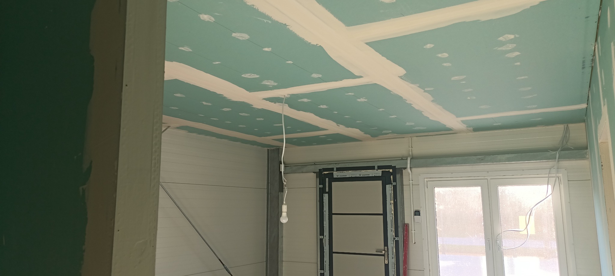 Niebieska płyta gipsowo-kartonowa na suficie z widocznymi łączeniami wypełnionymi masą szpachlową, surowa żarówka zwisająca na kablu, widoczne elementy metalowej konstrukcji i fragment ściany...