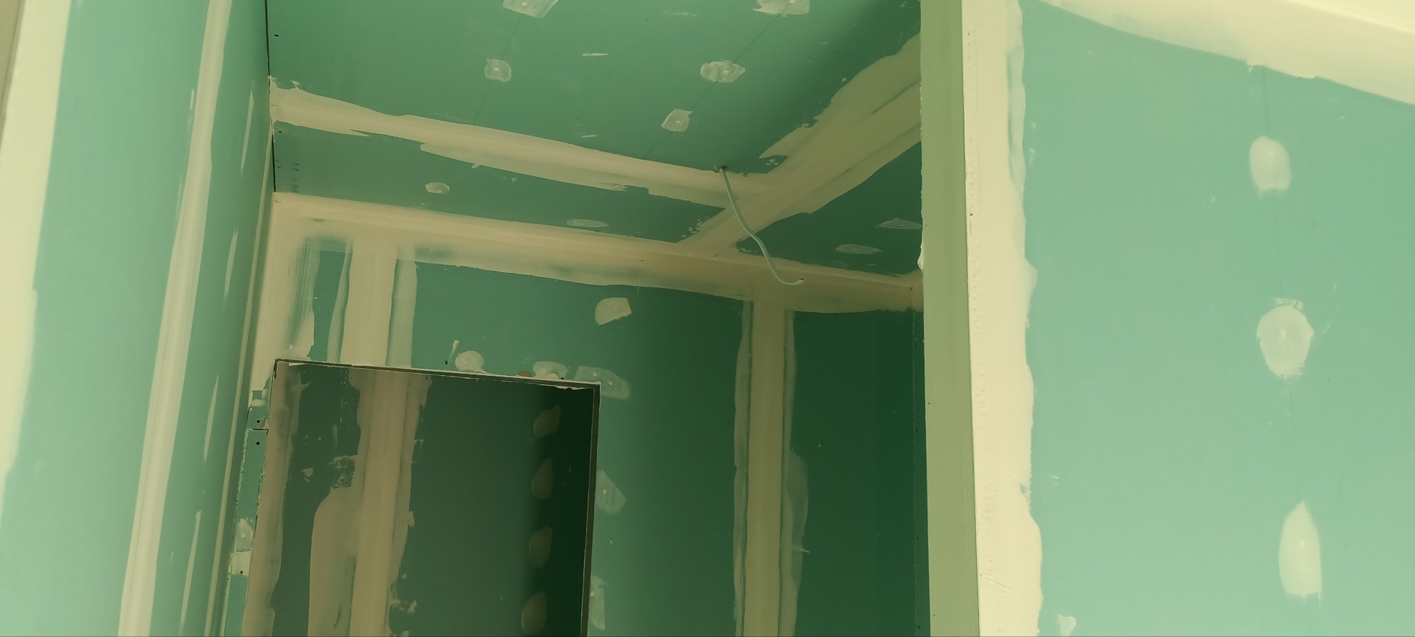 Wnętrze w trakcie prac wykończeniowych: ściany i sufit pokryte płytami kartonowo-gipsowymi, spoiny zaszpachlowane, widoczne otwory na oświetlenie w suficie, wystający kabel.