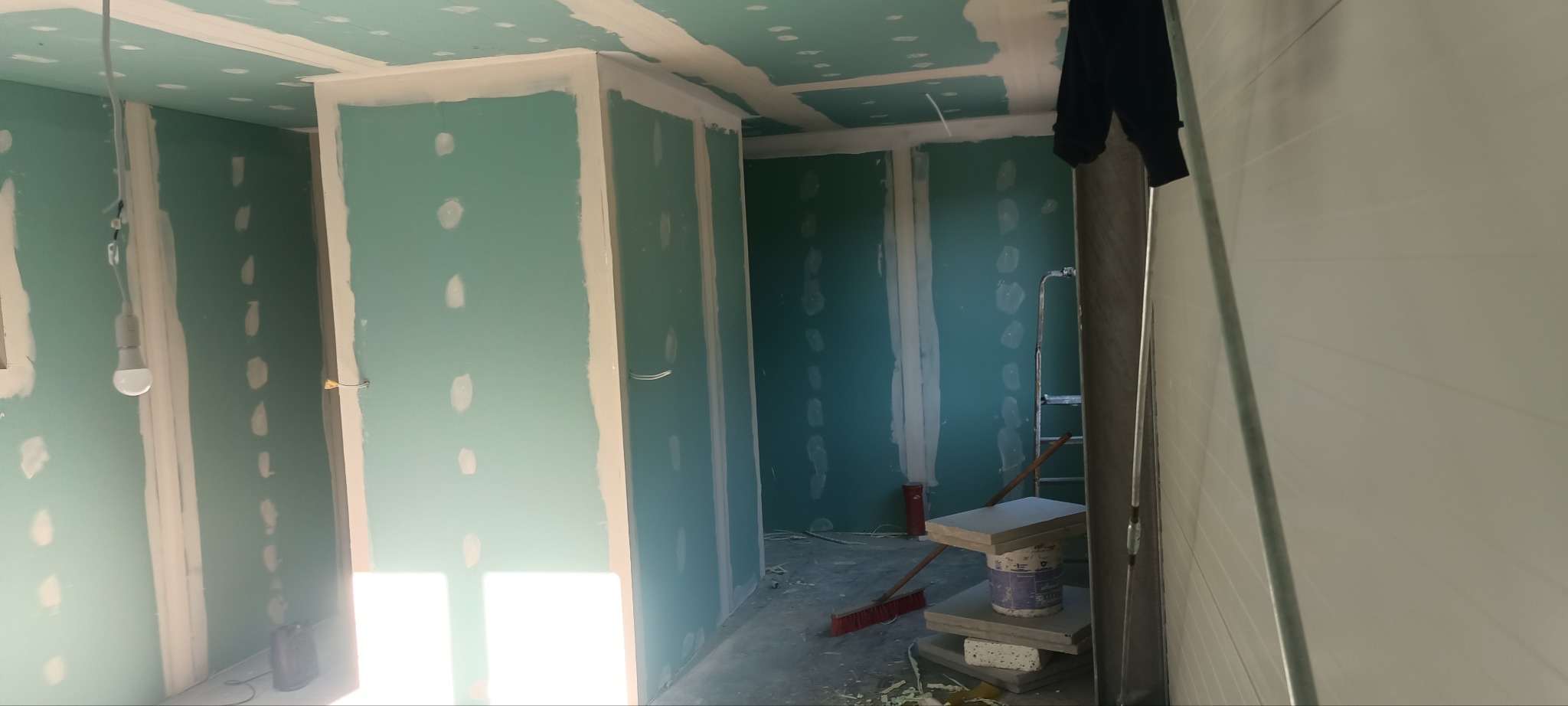 Wnętrze w trakcie prac wykończeniowych: zielone płyty gipsowo-kartonowe na ścianach i suficie, widoczne łączenia szpachlowane, wisząca żarówka, drabina i narzędzia budowlane w tle.