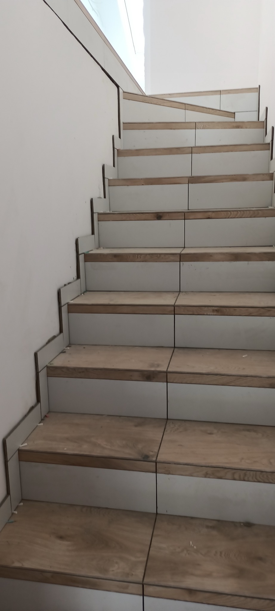 Schody w trakcie wykańczania z płytkami imitującymi drewno i białymi podstopnicami, widoczne krzyżyki dystansowe.