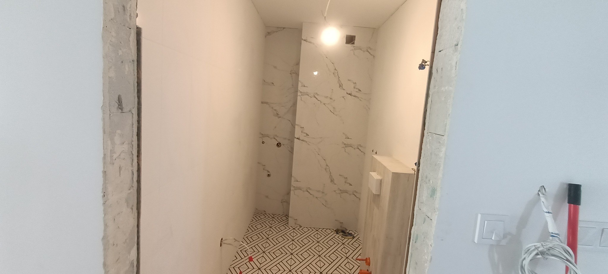 Wnętrze łazienki w trakcie prac wykończeniowych: ułożone płytki podłogowe z geometrycznym wzorem, marmurowe płytki ścienne, surowy betonowy otwór drzwiowy, widoczne instalacje elektryczne.