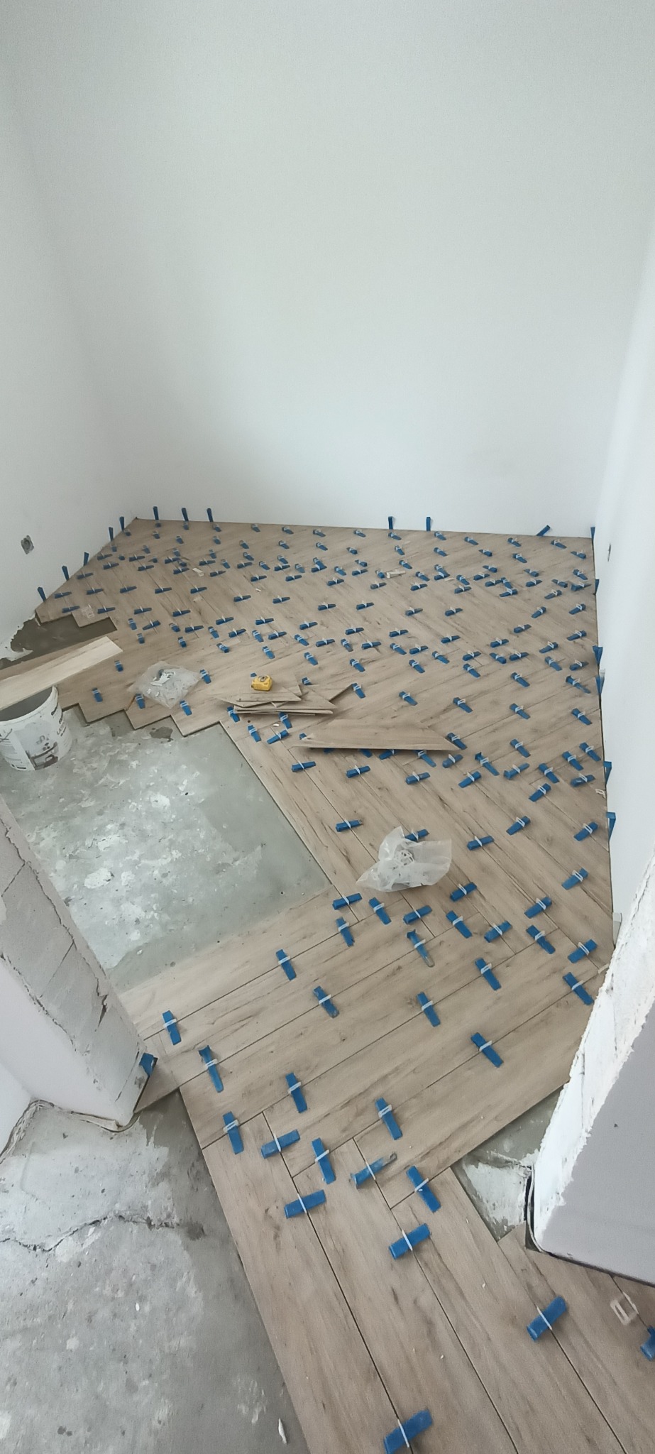 Układanie paneli podłogowych imitujących drewno w jodełkę, z użyciem niebieskich klipsów poziomujących, widoczny fragment surowej podłogi i wiadro z zaprawą.