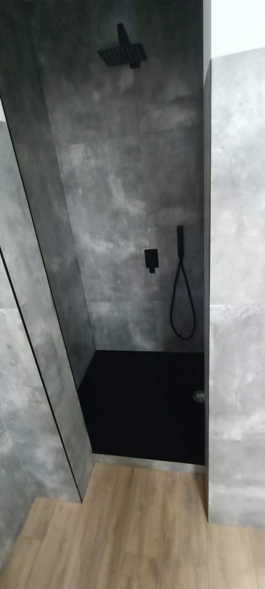 Nowoczesna kabina prysznicowa z szarymi płytkami imitującymi beton, czarną armaturą i czarnym brodzikiem, widok z boku, na podłodze panele podłogowe imitujące drewno.