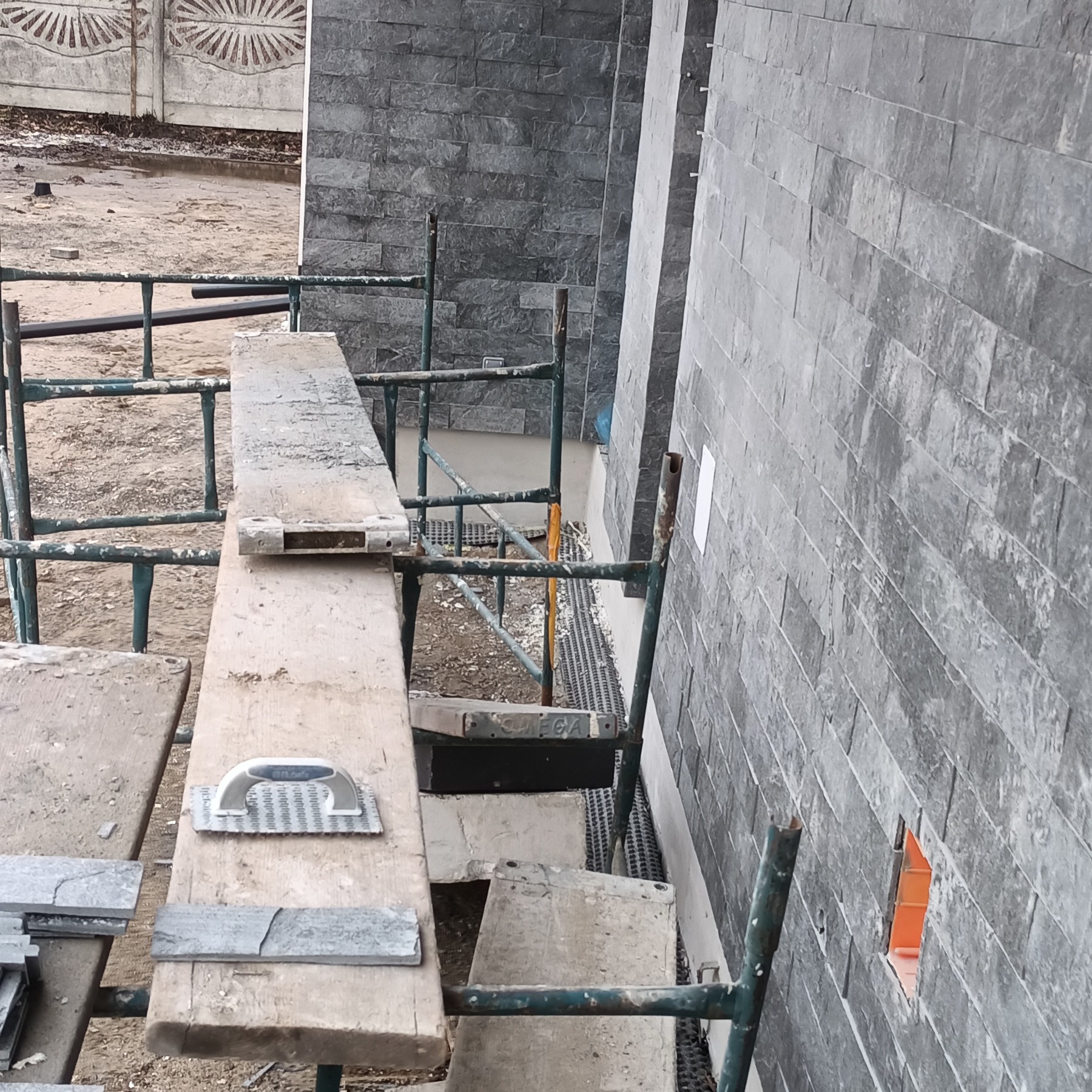 Montaż szarej okładziny kamiennej na elewacji budynku, widok ze rusztowania z narzędziami i fragmentami płytek.