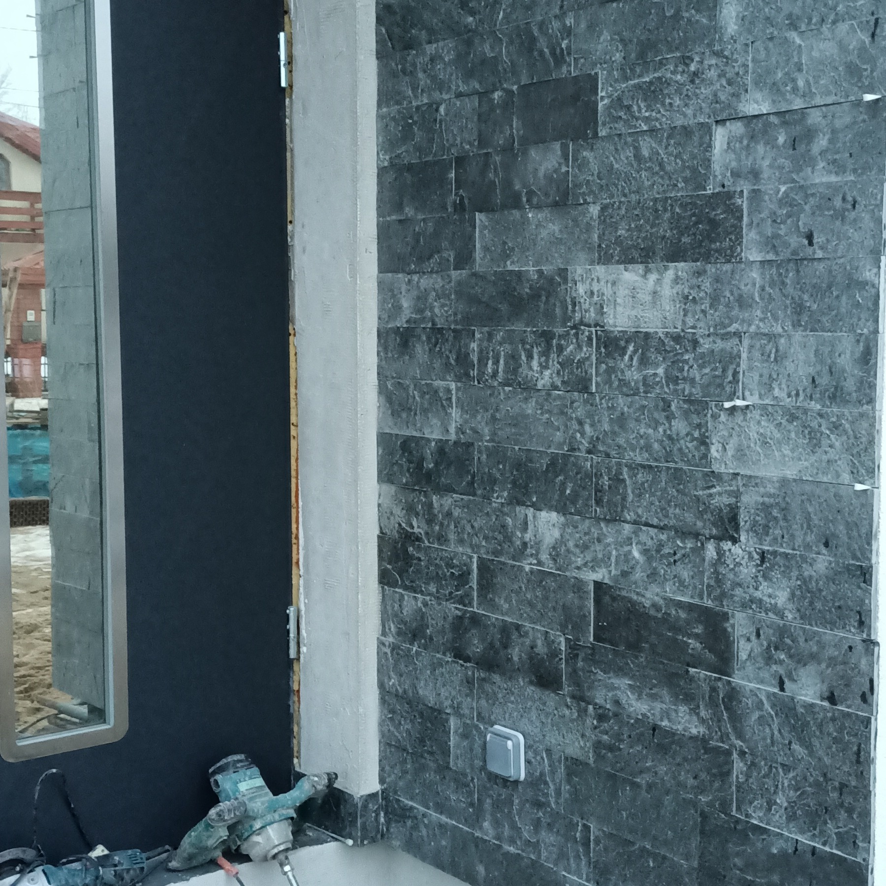 Ściana wyłożona szarym kamieniem dekoracyjnym, widoczna puszka elektryczna, obok stoją narzędzia budowlane: młot udarowy i wiertarka.