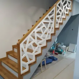 Jasne drewniane schody z białą, geometryczną balustradą w nowoczesnym domu, widoczne narzędzia i materiały budowlane w tle.