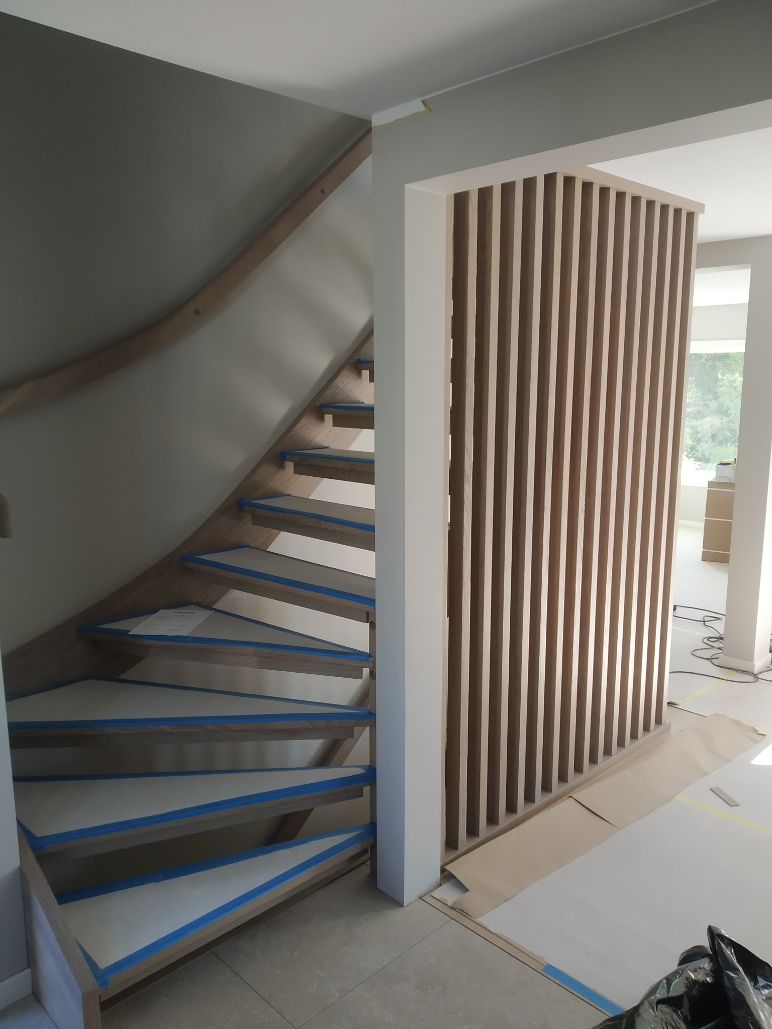 Wnętrze w trakcie wykończenia: drewniane schody z zabezpieczeniem taśmą malarską na stopniach oraz ażurowa ścianka działowa z drewnianych listew.