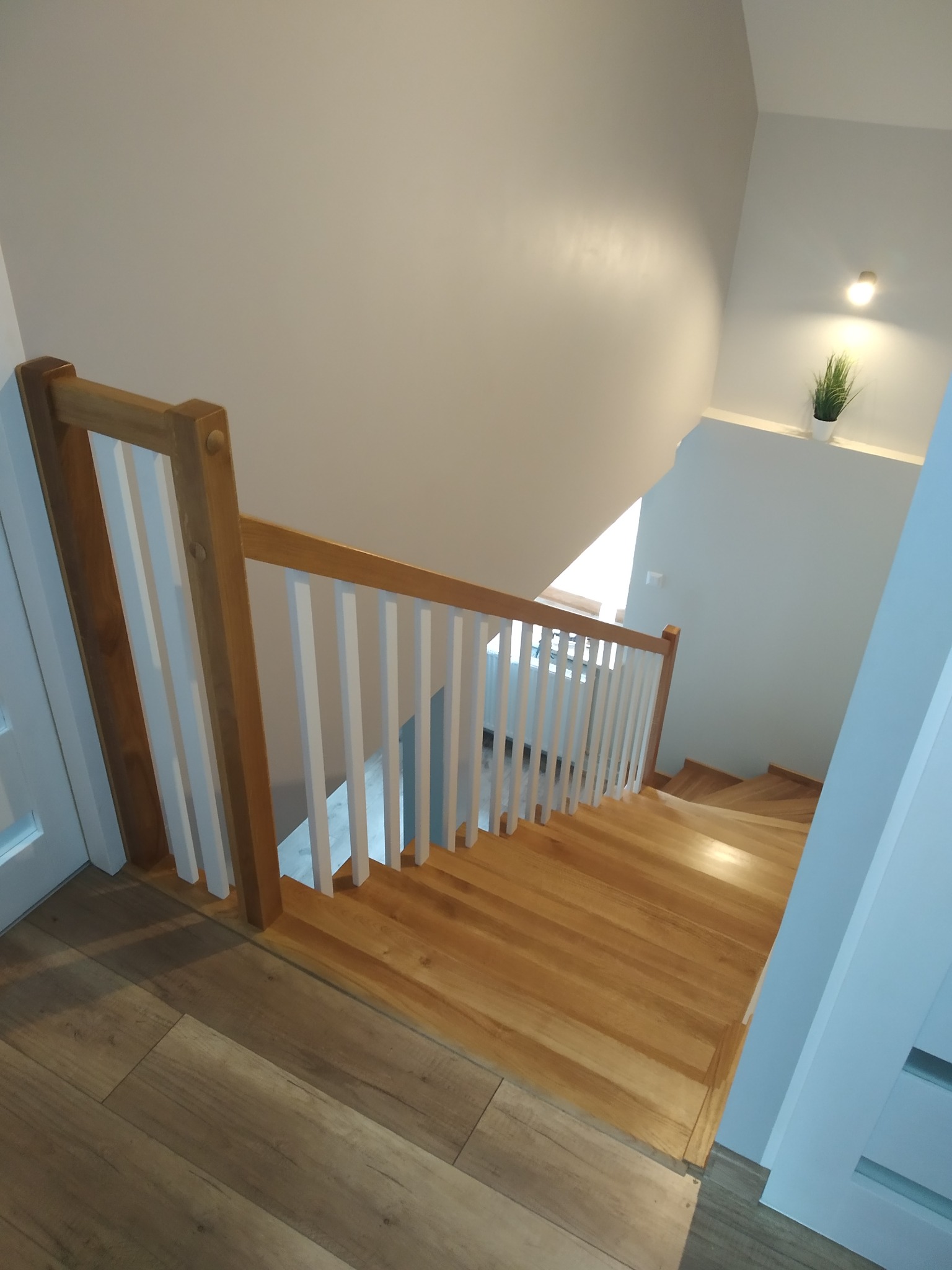 Jasne, drewniane schody z białą balustradą w nowoczesnym wnętrzu, widok z góry, z delikatnym oświetleniem i rośliną w tle.